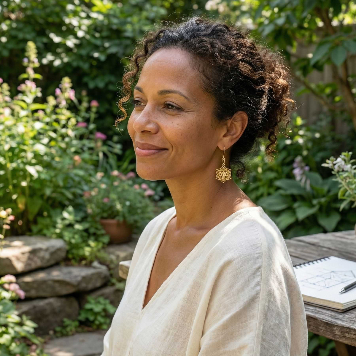 Woman sitting outdoors in a garden setting with flowers and plants around her wearing Mandala earrings.