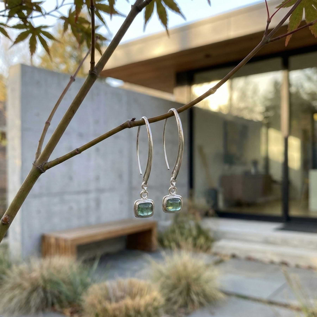 Silver drop earrings with green tourmaline cabochons hanging from a branch with a modern building in the background
