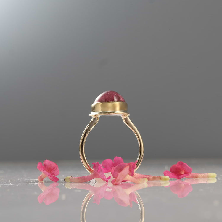Gold ring with a red gemstone on a reflective surface with pink flowers.