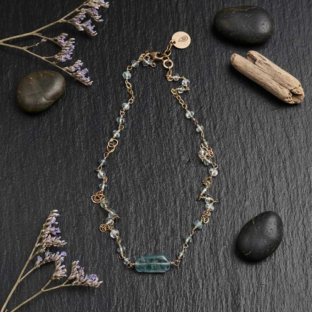 Flat lay of the Helena necklace featuring a blue aquamarine bar pendant and dainty faceted beads wire-wrapped into a 14k gold filled chain on a dark slate background with stones and flowers.