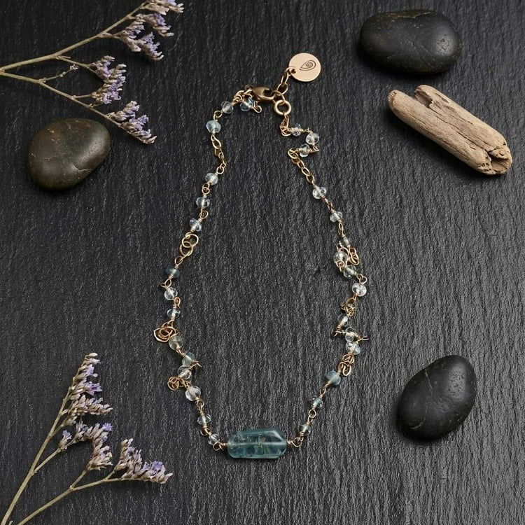Flat lay of the Helena necklace featuring a blue aquamarine bar pendant and dainty faceted beads wire-wrapped into a 14k gold filled chain on a dark slate background with stones and flowers.