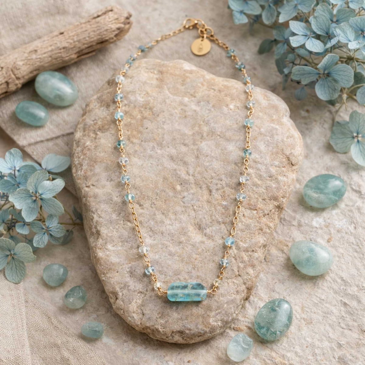 Aquamarine Bar Gold Necklace with blue beads on a stone surrounded by green stones and leaves.