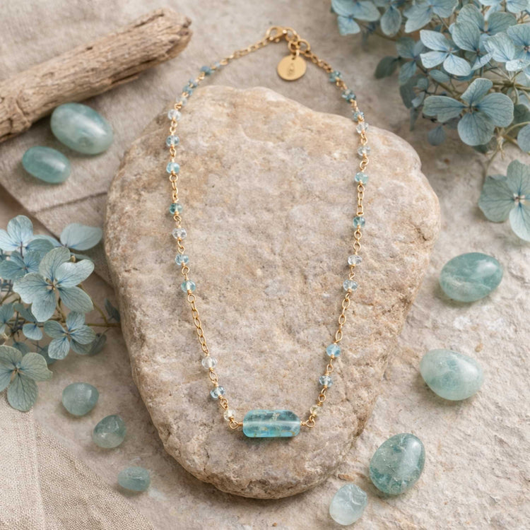 Aquamarine Bar Gold Necklace with blue beads on a stone surrounded by green stones and leaves.