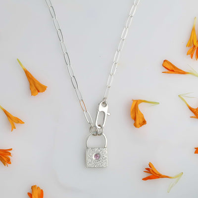 Tiny Argentium silver padlock pendant with flush-set pink sapphire on sterling silver paperclip chain, styled with orange flower petals.