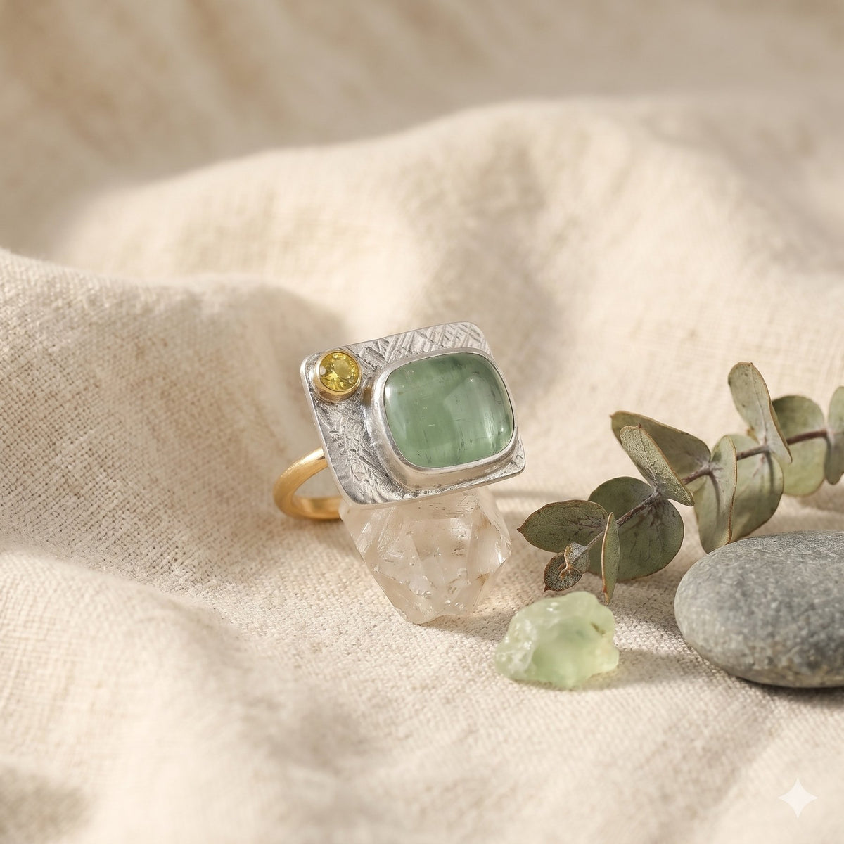 Silver ring with green stone and yellow gemstone on a textured surface with eucalyptus leaves and stones.