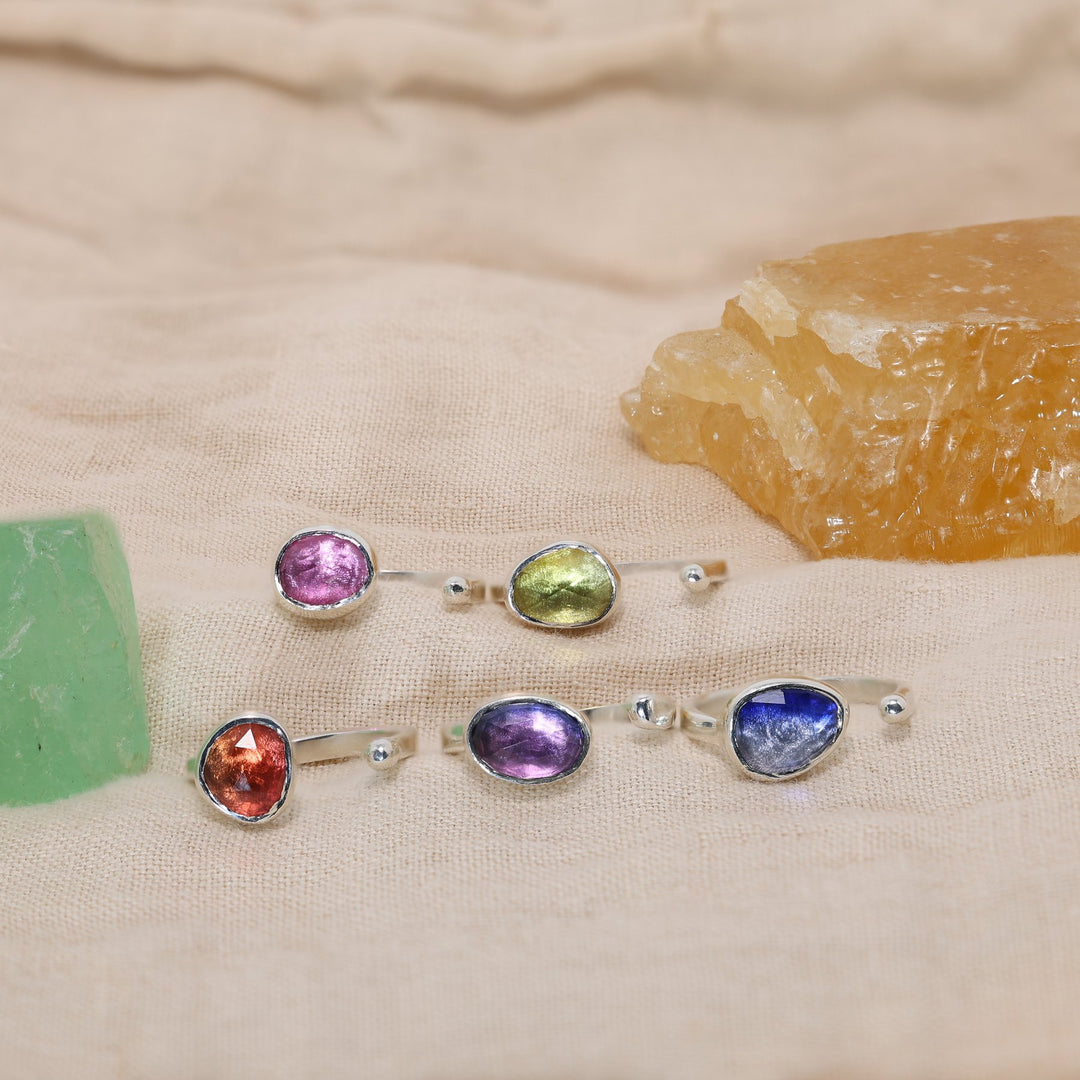 Set of five silver rings with colorful gemstones on a textured beige surface.