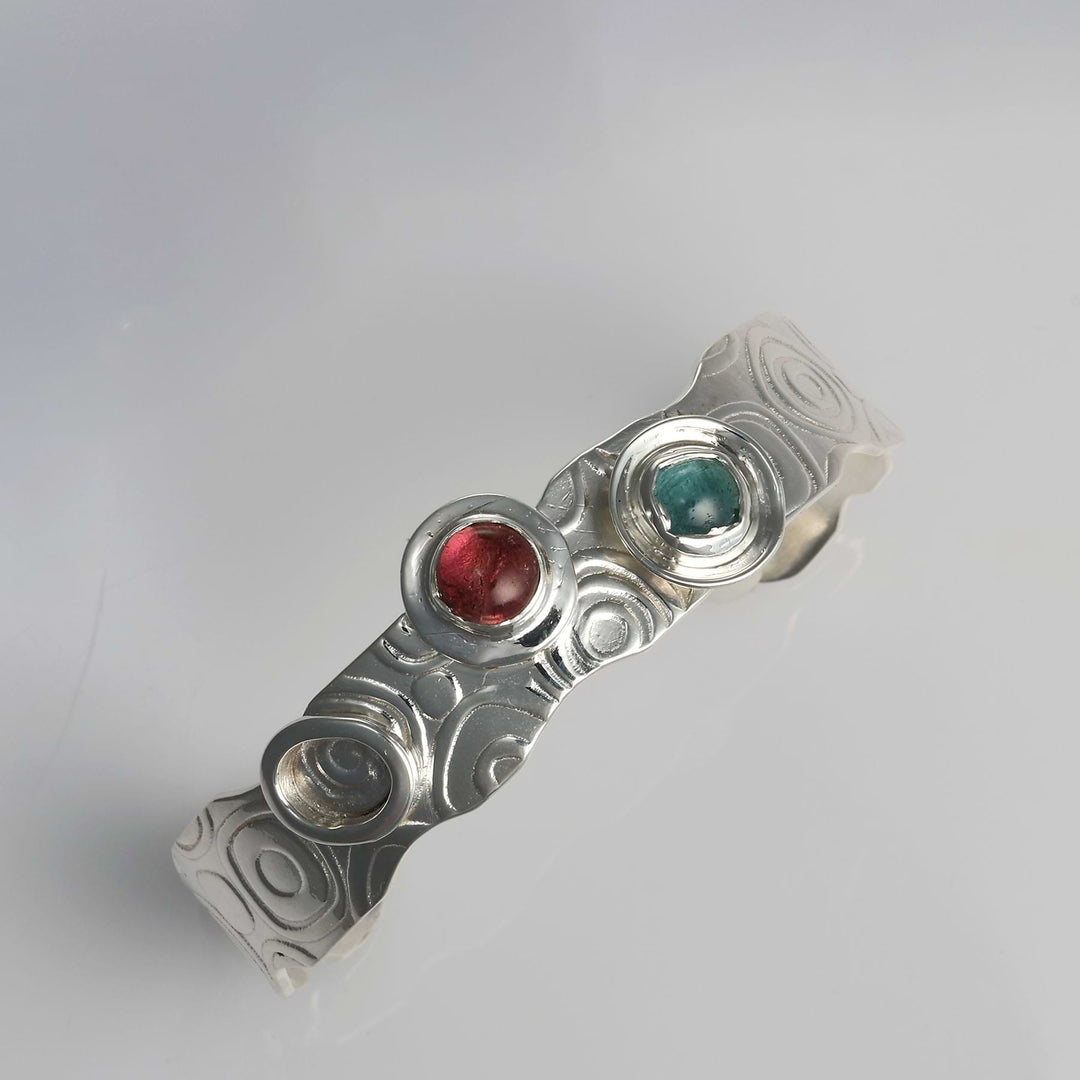 Top down view on textured Argentium silver cuff bracelet featuring retro swirl patterns and bezel-set multi-color tourmalines.